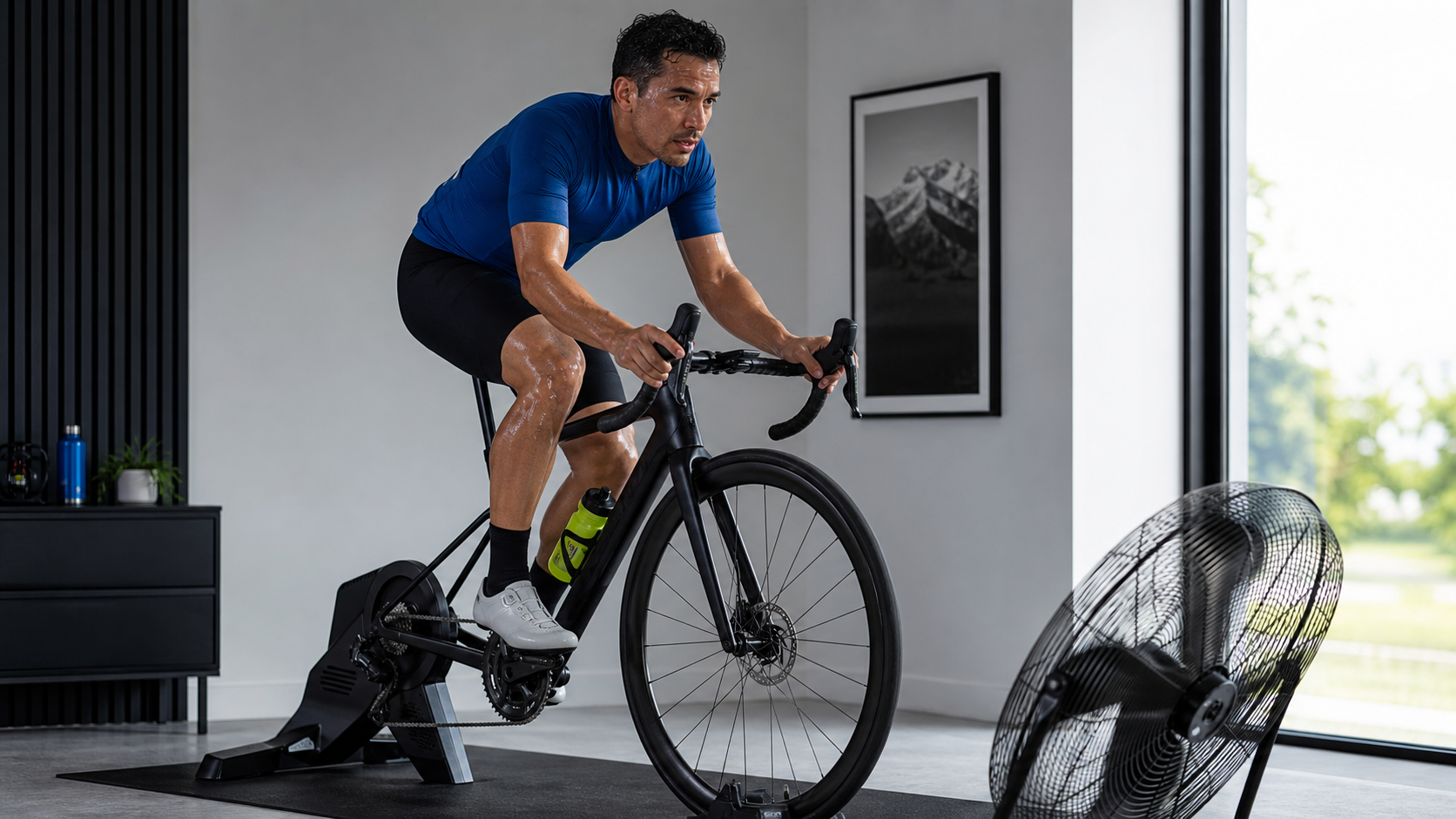 Latino man in a royal-blue cycling top riding an indoor trainer beside a large floor fan in a clean modern training room