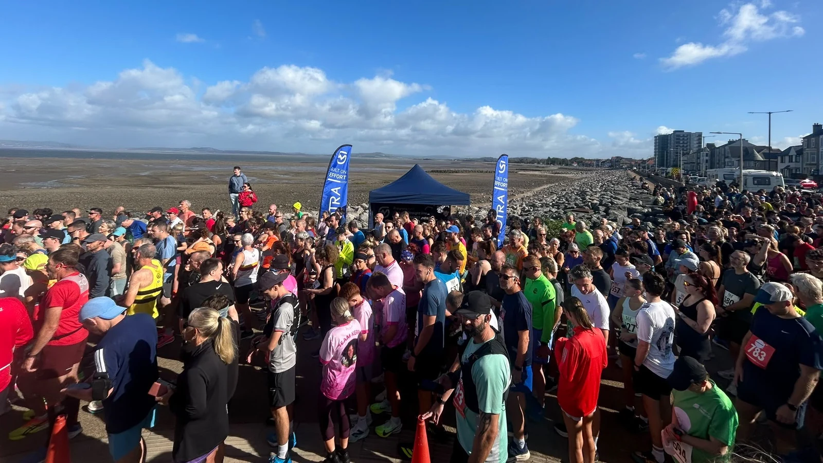 Runners gathering in front of the blue MOTRA stand and flags beside Morecambe Bay.