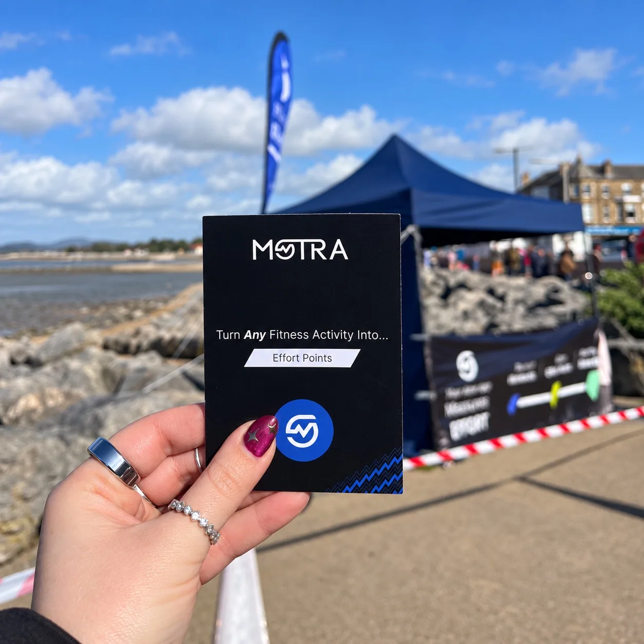 A black MOTRA Effort Points card held in front of the blue event stand on the Morecambe promenade.