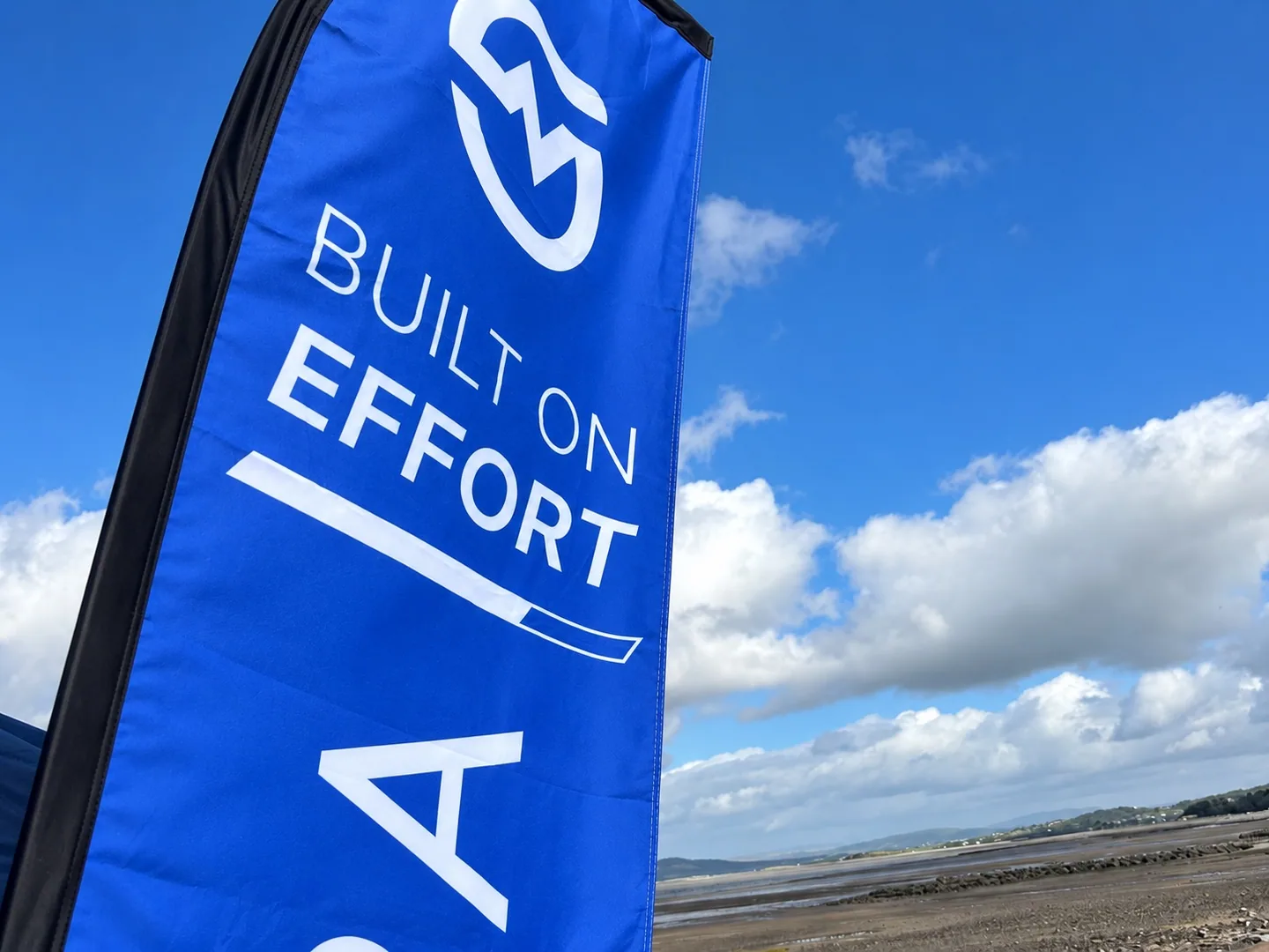 A blue MOTRA Built on Effort flag against the sky and Morecambe Bay.