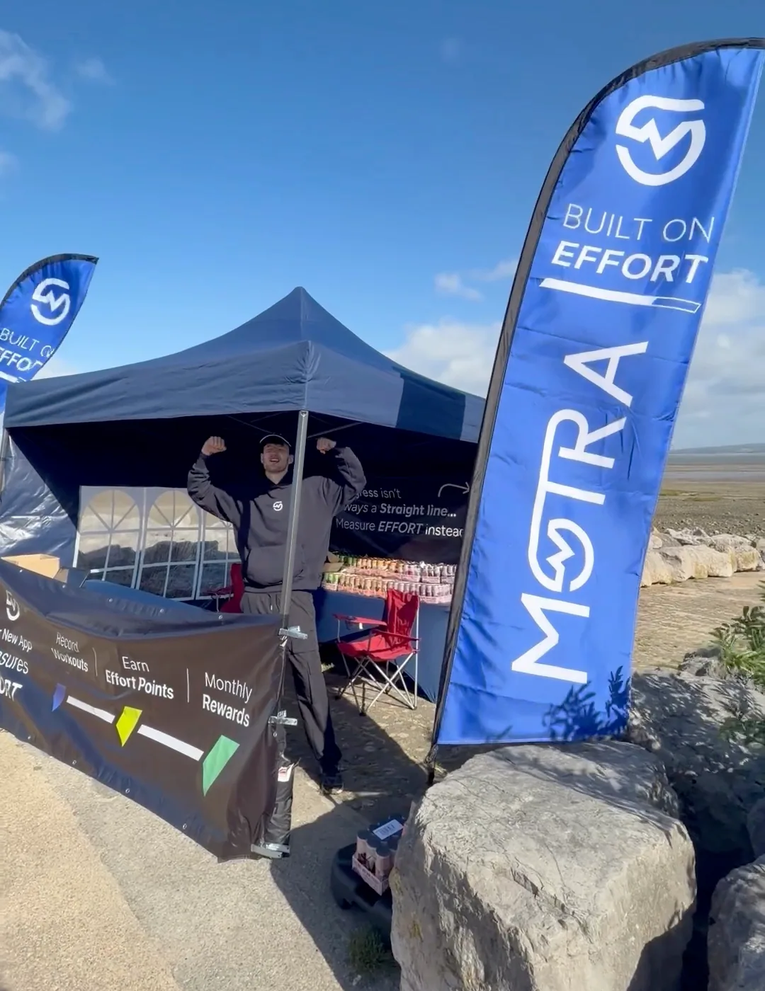 A MOTRA team member raising his arms beside the blue Built on Effort flag at the seafront stand.