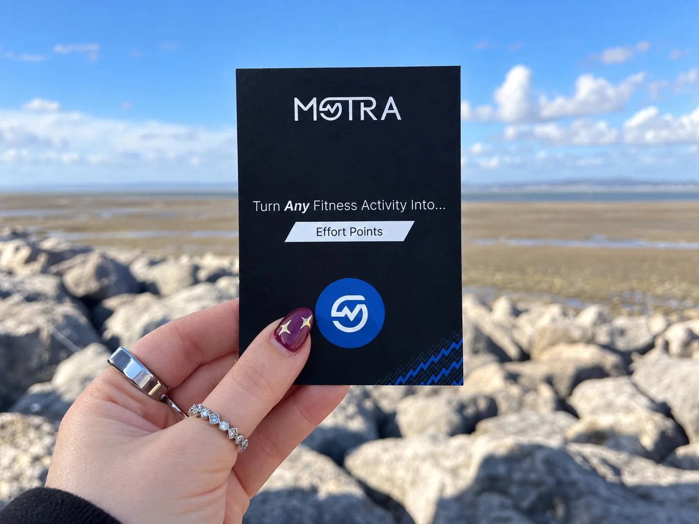 A hand holding a black MOTRA Effort Points card above the rocks beside Morecambe Bay.