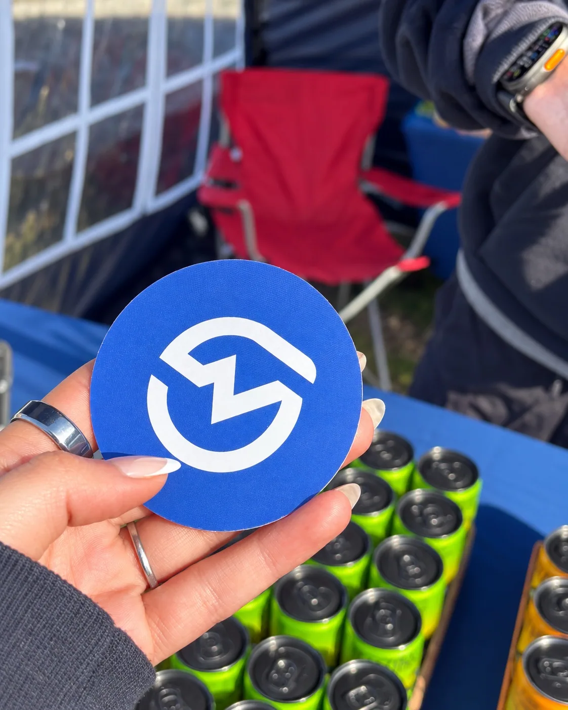 A round blue MOTRA logo card held above green and orange TENZING cans at the event stand.