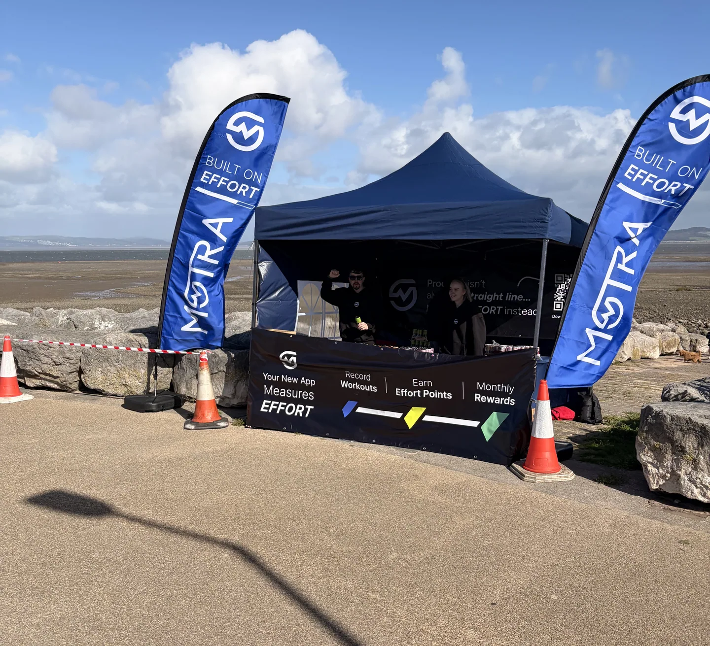 The MOTRA event stand between two blue Built on Effort flags, with team members and Morecambe Bay behind it.