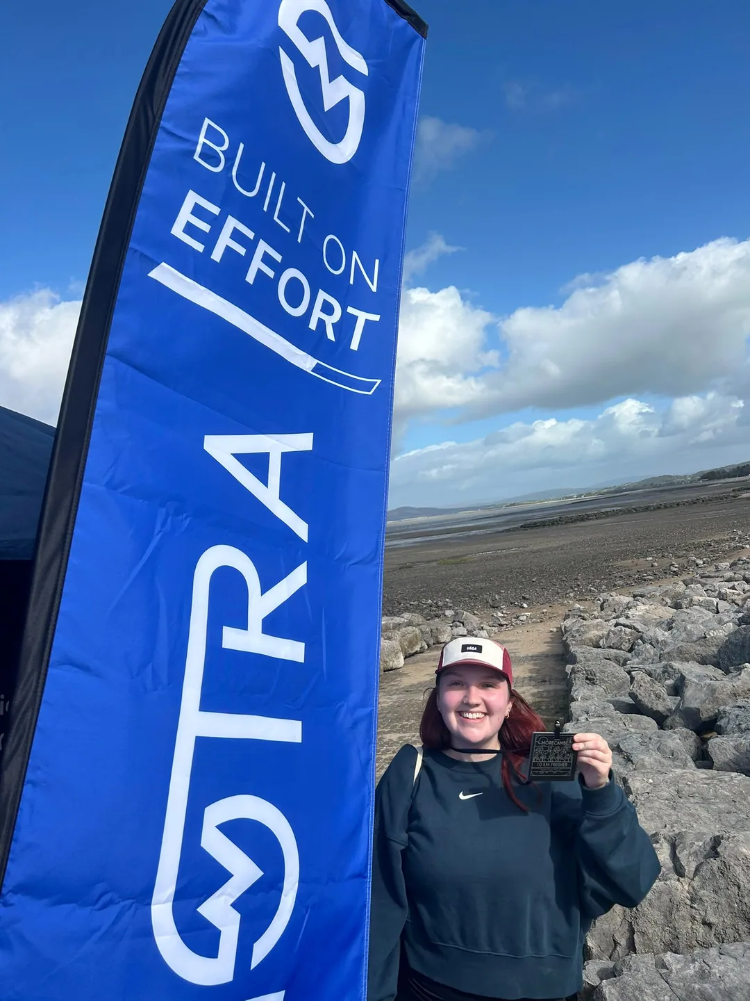 A smiling 10K finisher holding a medal beside a blue MOTRA Built on Effort flag at Morecambe Bay.