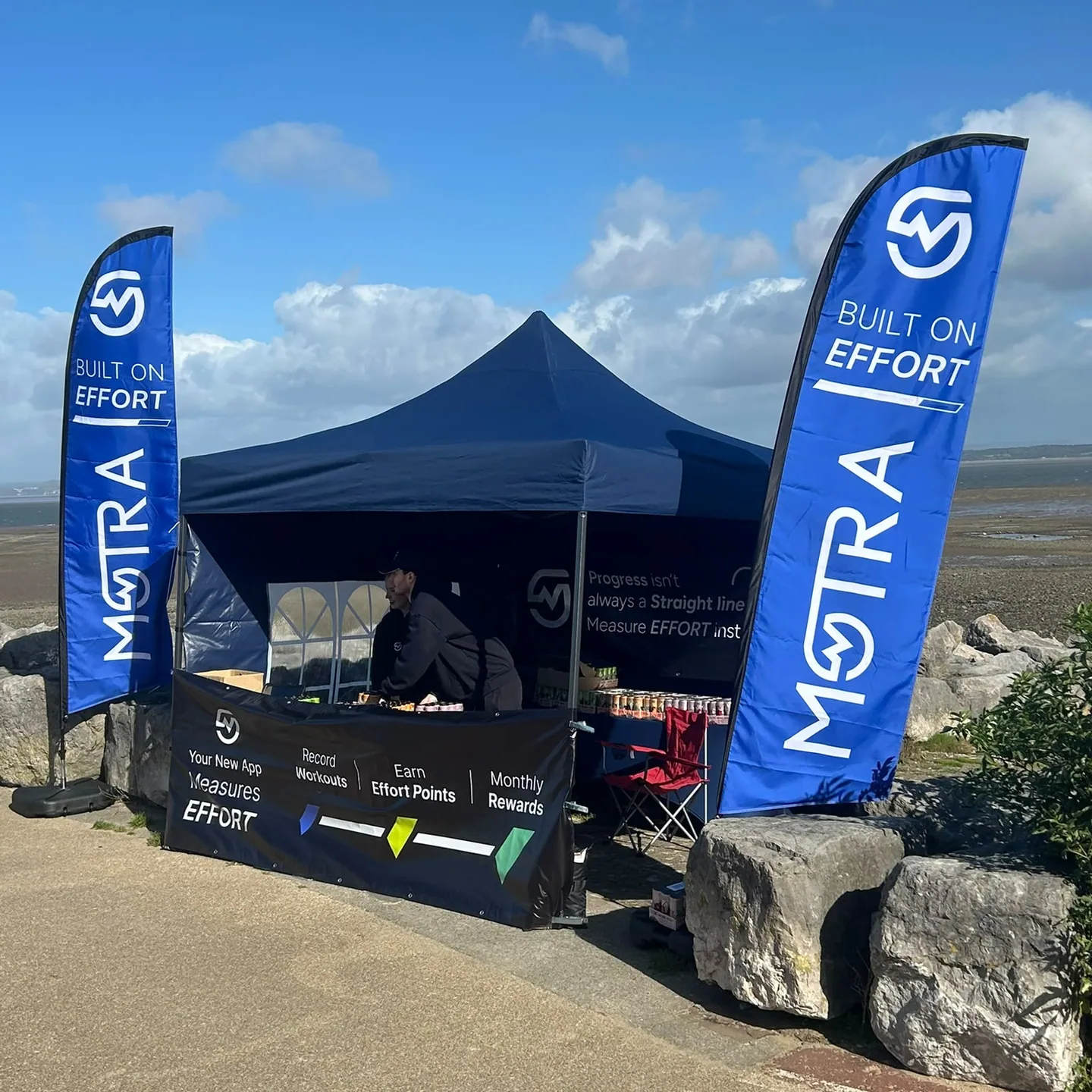 A MOTRA team member at the blue event stand between two Built on Effort flags beside Morecambe Bay.