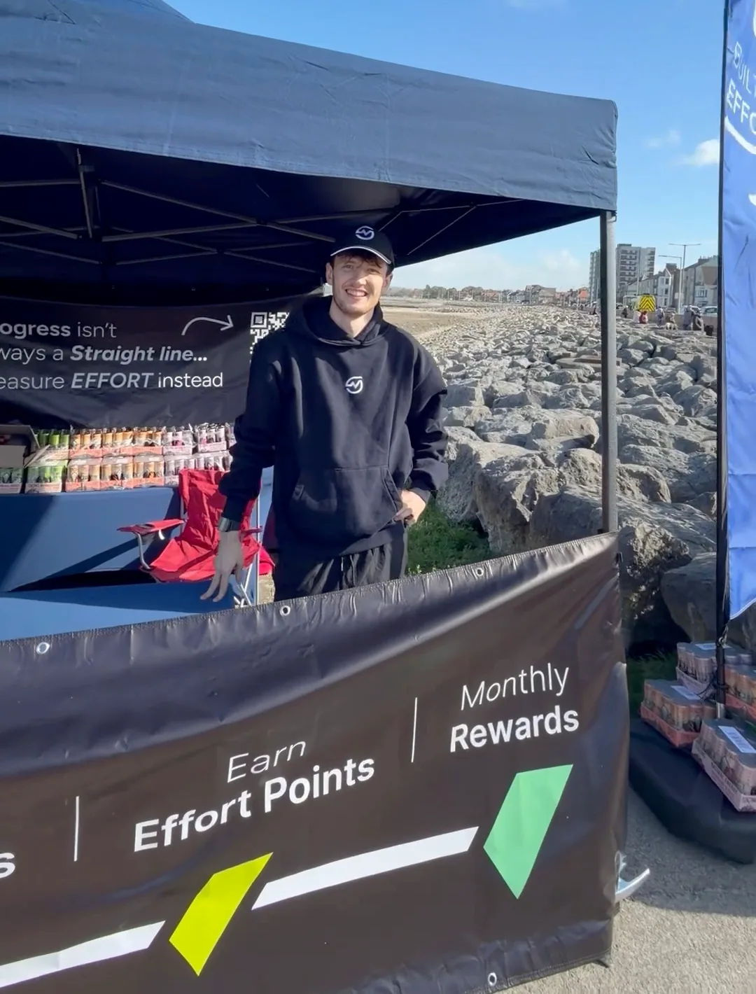 A MOTRA team member standing inside the event stand with TENZING cans and the bay behind him.
