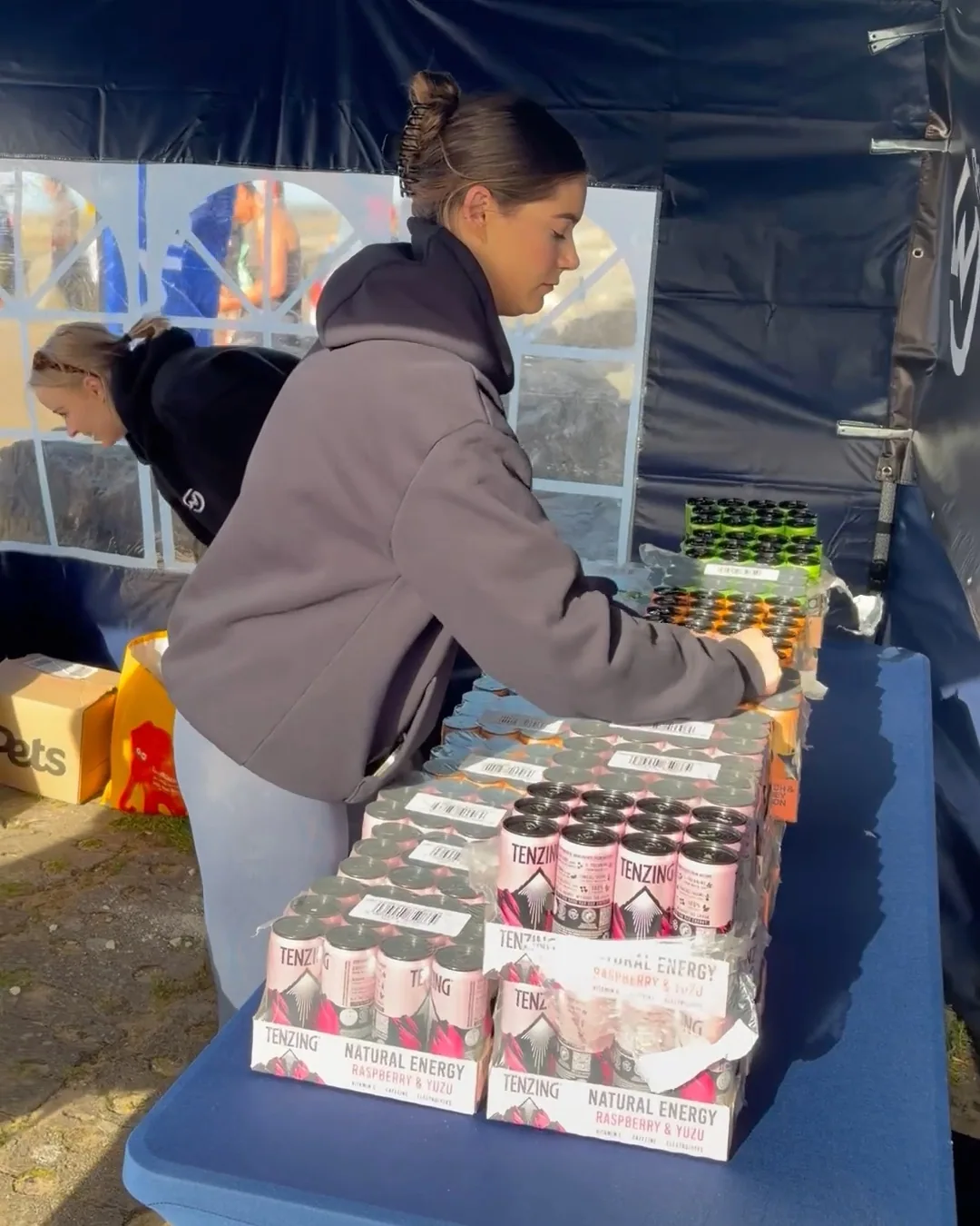 A MOTRA volunteer arranging trays of TENZING cans on the blue drinks table beneath the canopy.