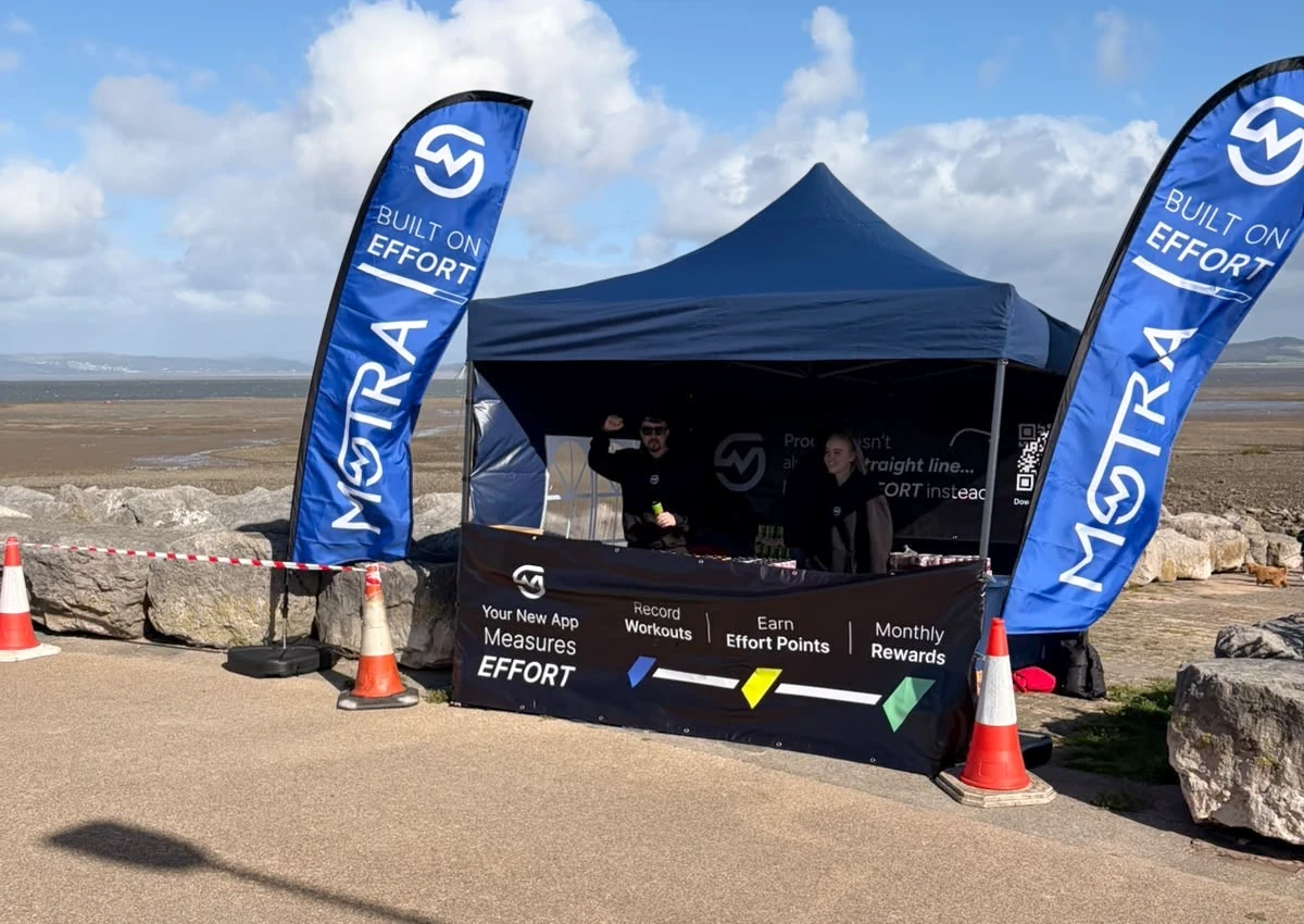 Two MOTRA team members behind the blue event stand between Built on Effort flags beside Morecambe Bay.