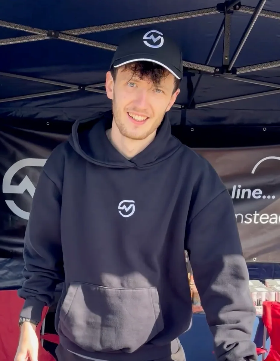 A smiling MOTRA volunteer wearing a black logo cap and hoodie beneath the event canopy.