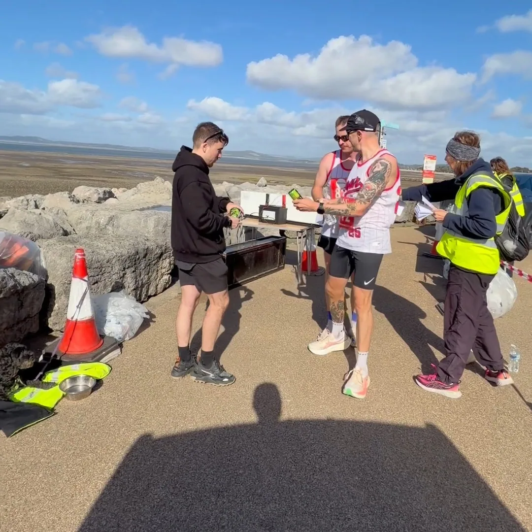 A MOTRA team member sharing green TENZING cans with runners beside a race marshal on the promenade.