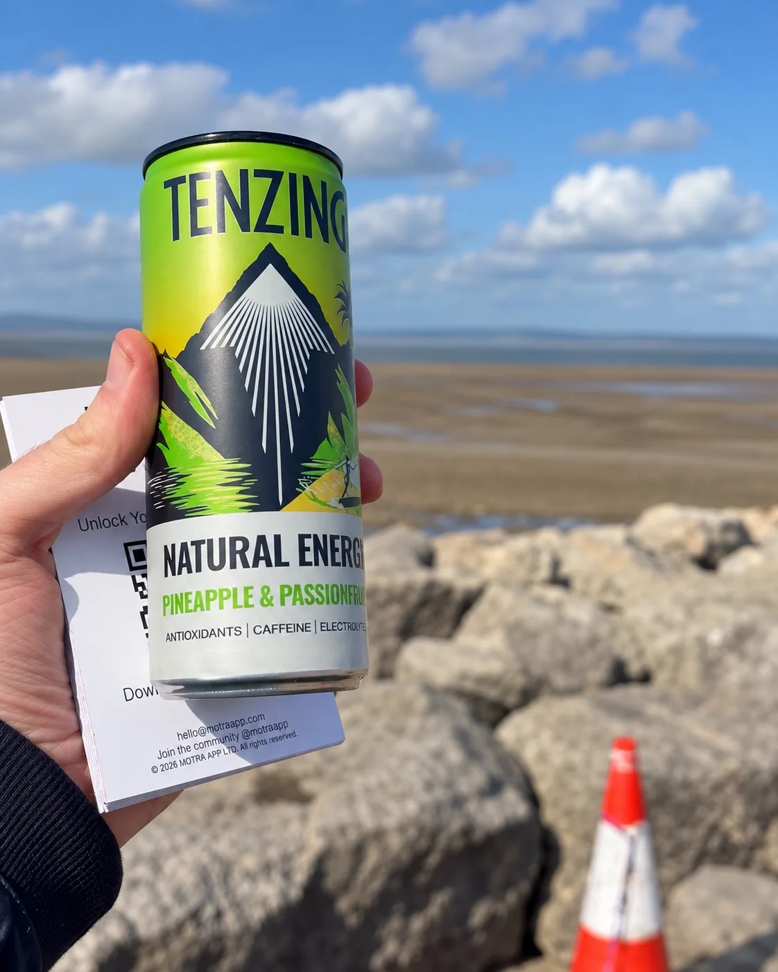A TENZING Pineapple & Passionfruit can and MOTRA QR card held in front of Morecambe Bay.