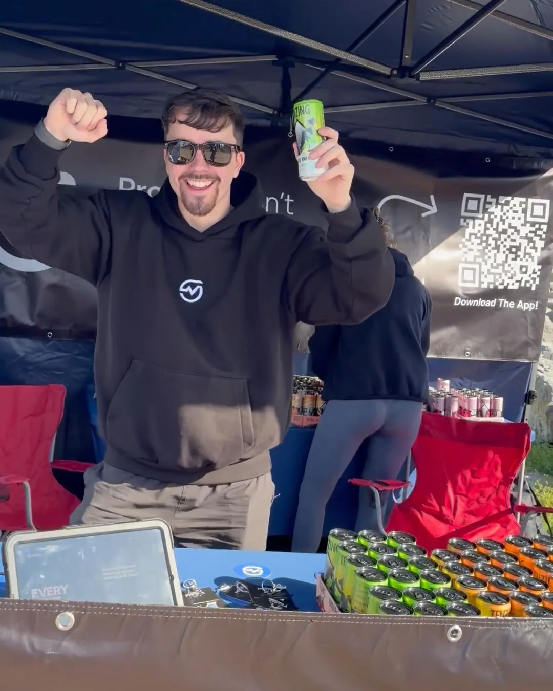 A smiling MOTRA team member raising a green TENZING can behind the event stand.