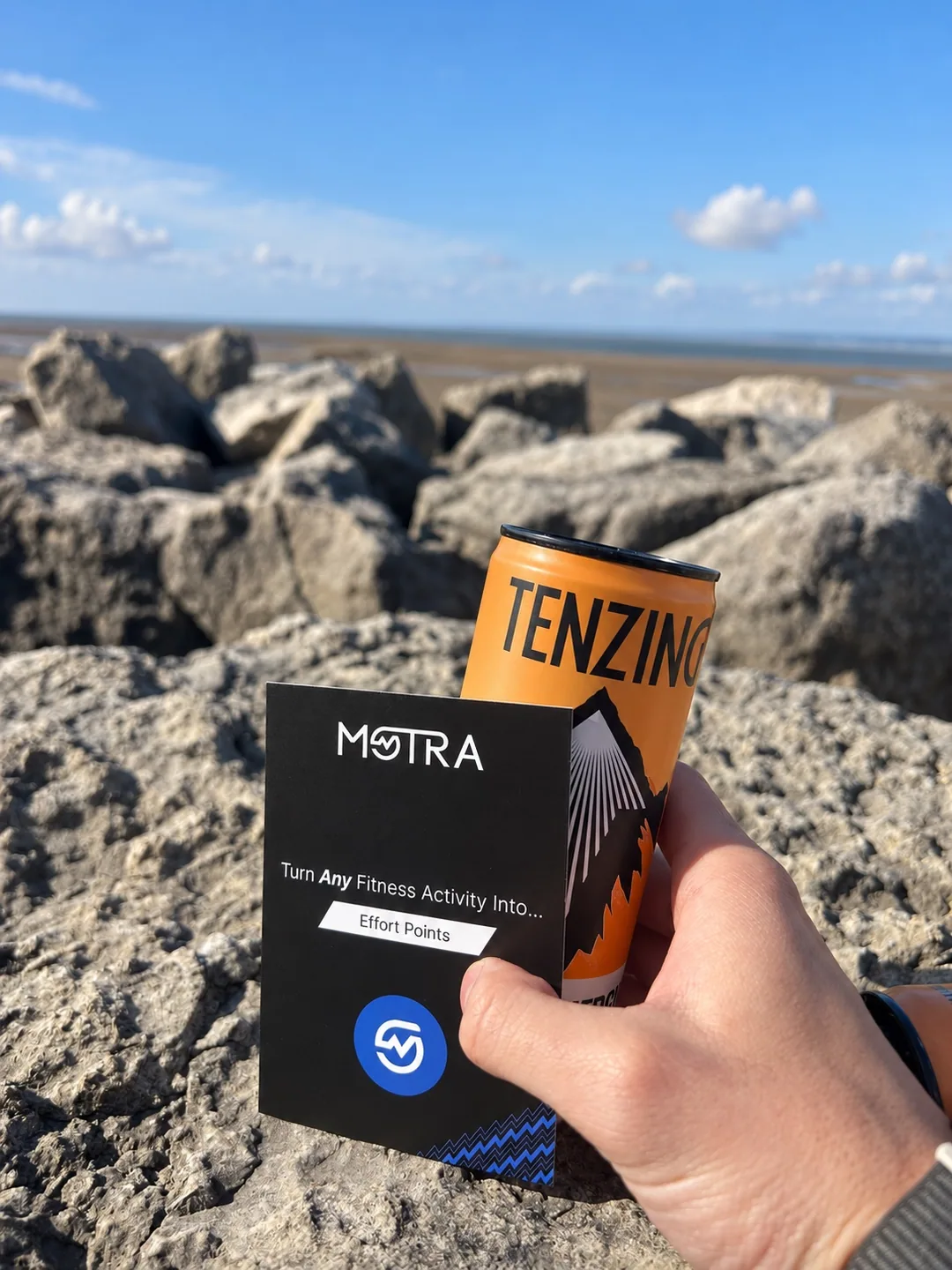 A hand holding an orange TENZING can and a MOTRA card beside the rocks at Morecambe Bay.