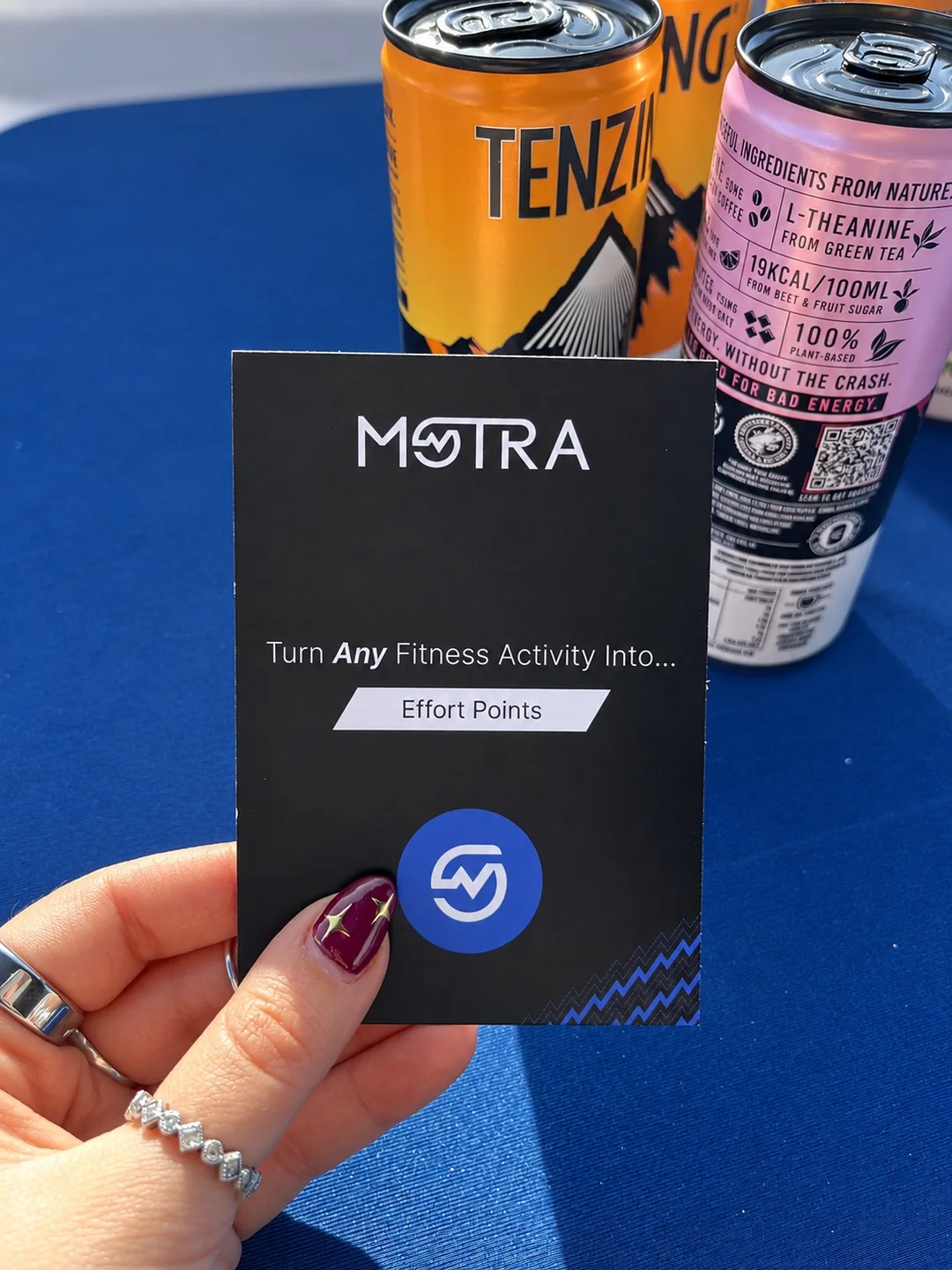 A hand holding a black MOTRA card in front of orange and pink TENZING cans on the stand’s blue table.