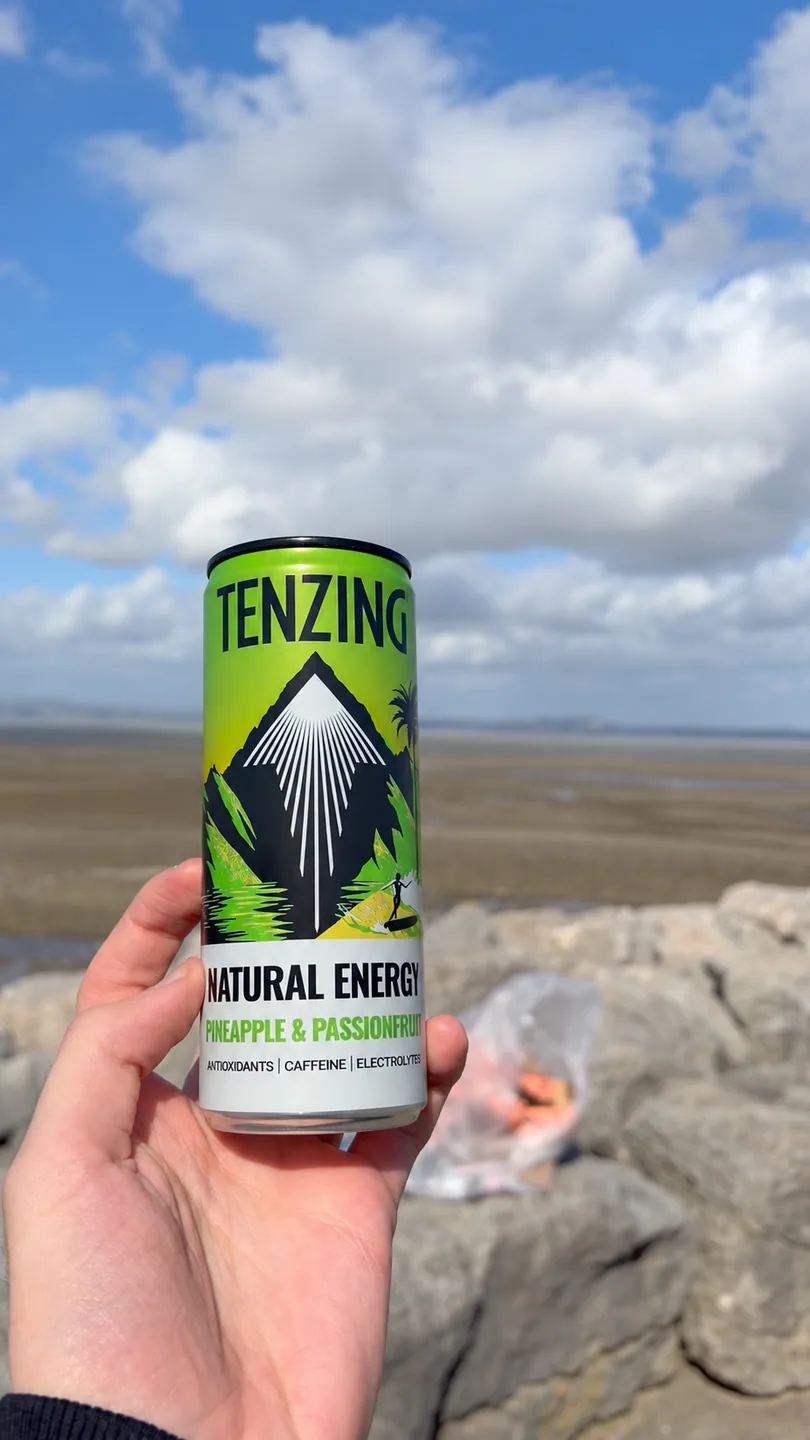 A TENZING Pineapple & Passionfruit can held above the seafront rocks with Morecambe Bay behind it.