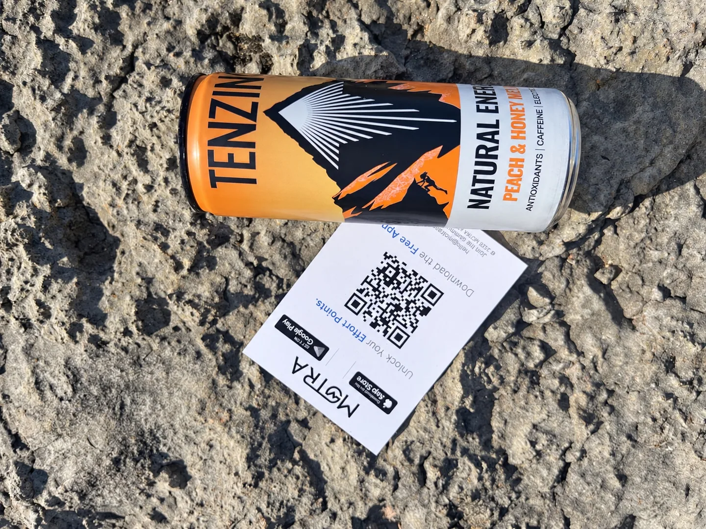 A TENZING Peach & Honey Melon can resting beside a MOTRA QR card on Morecambe’s seafront rocks.