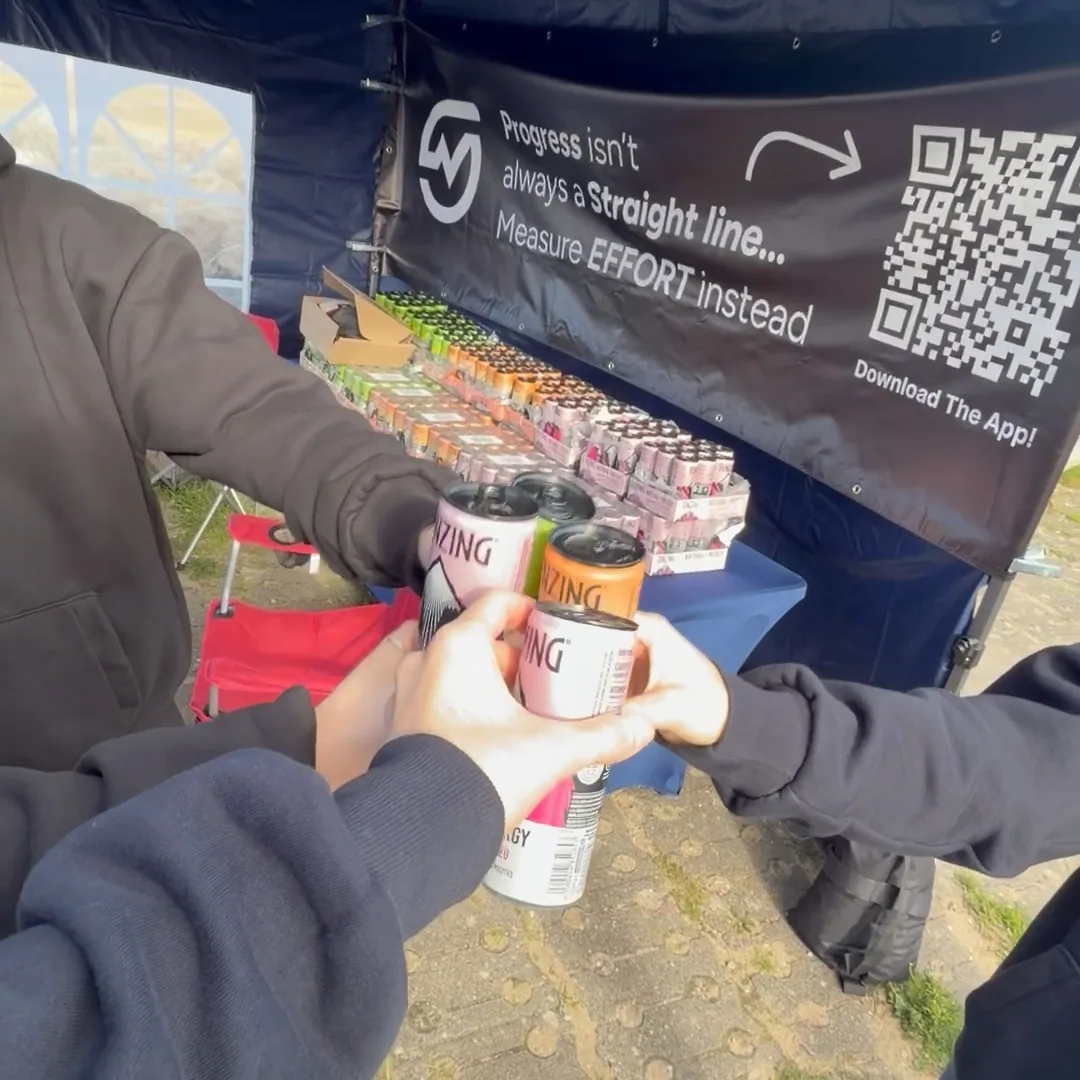 Hands raising TENZING cans together in front of the MOTRA banner and drinks table.