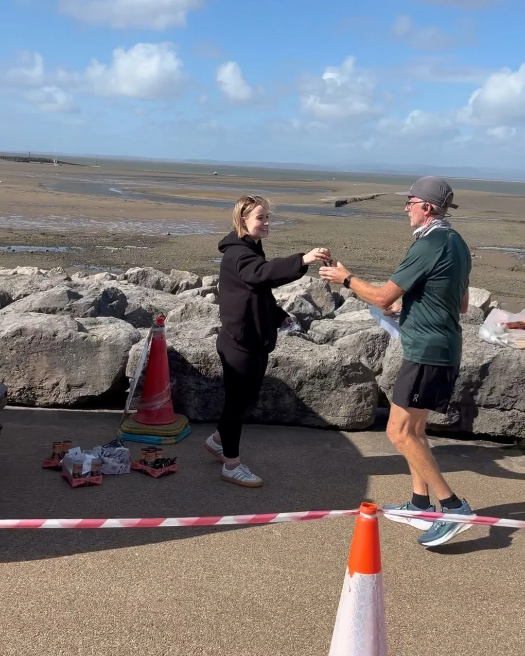 A MOTRA team member handing a TENZING can to a runner beside the rocks at Morecambe Bay.