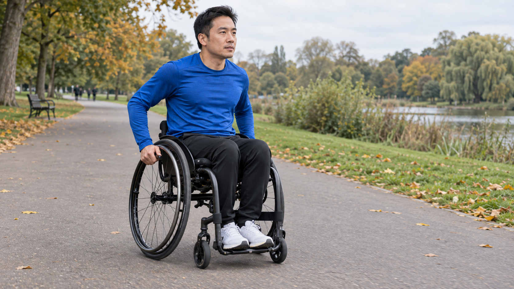 Man in a royal-blue training top propelling a manual wheelchair along a broad, smooth park path in early autumn.