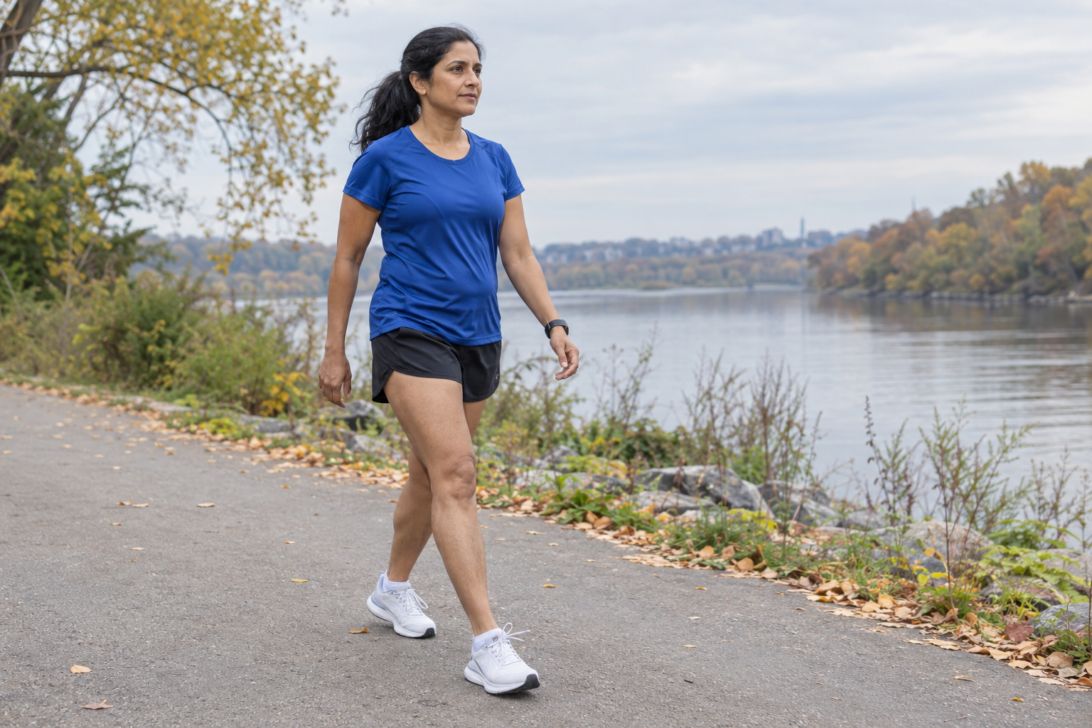 Woman in a royal-blue running shirt taking a walking break on a riverside training path in early autumn.